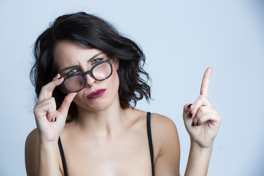 Ritratto di ragazza bianca con i capelli scuri e con occhiali da vista, alza l'indice con espressione severa , isolata su sfondo bianco