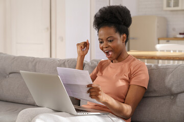 Excited millennial African American woman sitting on couch at home, holding and reading paper...