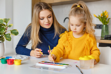 Mother and daughter painting at home. Cute little kid in yellow sweater having fun with parent and paints. Concept of early childhood education, hobby, talent, preschool leisure and parenting