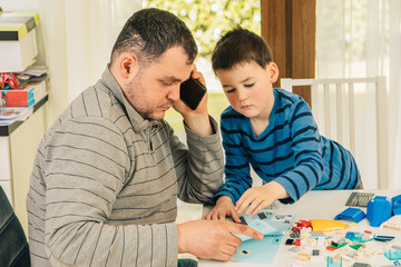 Boy playing with his father in the kitchen. Child builds something out of small parts. Father son relationship. Father is on the phone while playing with his child 
