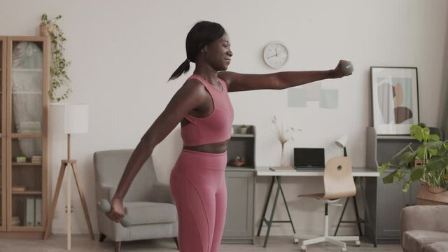 Medium shot of young African-American sportswoman wearing fashionable pink sportswear exercising with dumbbells in living room