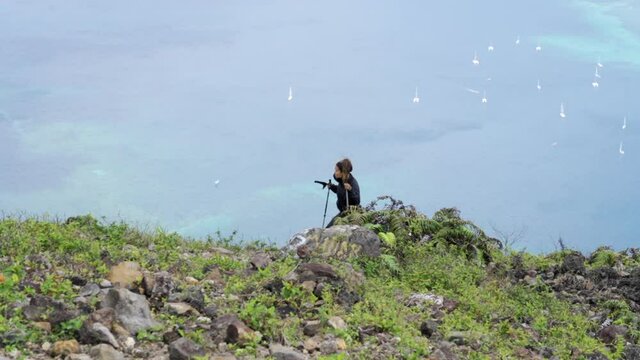 A striking slow motion of a young woman hiking up a steep green hill before a distant bay with tiny white sailboats - Banda, Indonesia