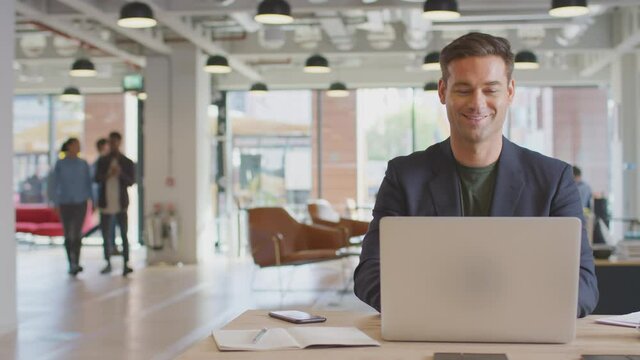Dolly shot of casually dressed businessman working on laptop taking coffee break at desk in modern open plan start up office with colleagues in background - shot in slow motion