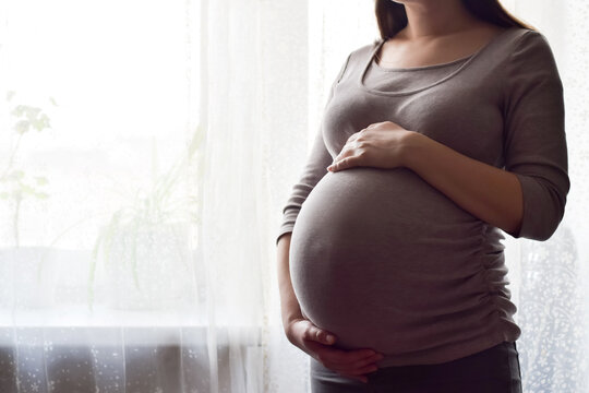 Pregnant woman with big belly at window. Maternity prenatal care and woman pregnancy concept. Young pregnant woman holds her hands on her swollen belly. Love concept. Horizontal, copy space.