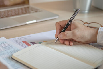 Young employee, businessman write note on notebook, analyzing charts on paperwork to  plan his spending for payday. Financial, finance person concept.