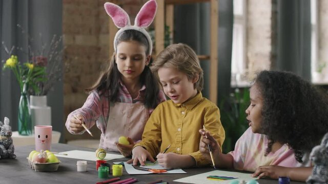 Handheld shot of cute girl wearing bunny ears headband painting on dyed Easter egg and talking to her friends making cards