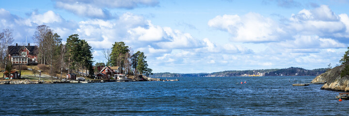 Panoramic view of the Baltic Sea Bay on sunny spring day. Rocky shores of Scandinavia covered with evergreen forests. Traditional Swedish red wooden houses on the coast. Beautiful white clouds on sky.