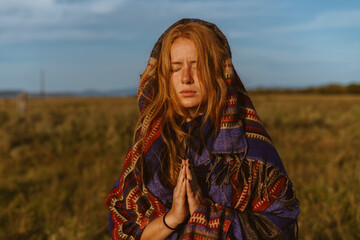 Against the background of the blue sky and steppe grass stands prayerfully folded hands red-haired girl with closed eyes with a cape on her head and shoulders. High quality photo