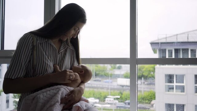 joyful lady silhouette holding little baby, gently touching and playing with funny kid while sitting near large window with city buildings outside
