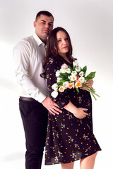 Husband and pregnant wife holding flowers on a white background. The guy holds the pregnant girl by the belly. Love and relationships on a white background.