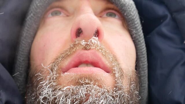 Close-up of the face of a bearded man standing in the cold outside in heavy snowfall and his beard is covered with hoarfrost