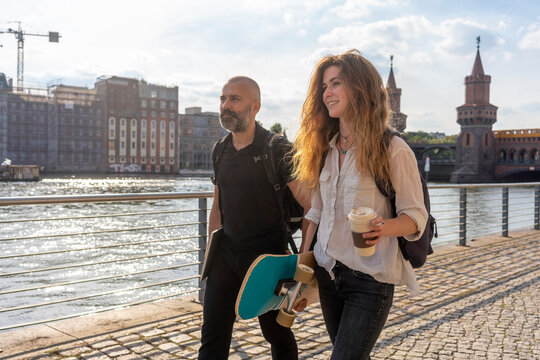 Man and female friend with skateboard on bridge, river, Oberbaum bridge and buildings in background, Berlin, Germany