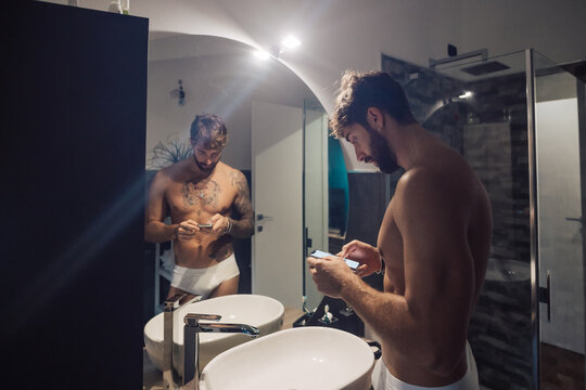 Mid adult man with tattoos using smartphone touchscreen at bathroom mirror, mirror image