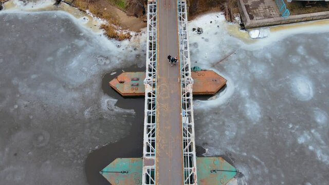 Top down view on unrecognizable people walking on steel pedestrian bridge through river with melting ice in winter. Concept of weekend and free time with active lifestyle in cold cloudy weather