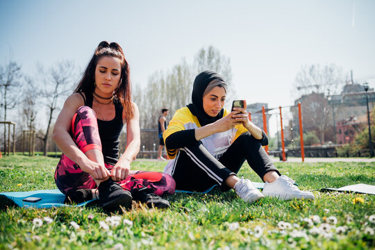 Calisthenics class at outdoor gym, young women sitting on grass tying laces and looking at smartphone