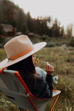 Young woman in stetson smelling wildflower on deck chair in rural valley, rear view, Mineral King, California, USA