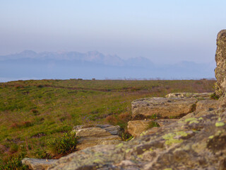 A panoramic view on an Alpine meadow in high mountains during an early morning. There is a fog covered high and massive mountain chain in the back. A pile of stones on the side.