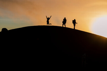 Silhouette of three climbers with their backpacks on top of the Acatenango volcano in Guatemala - Friends hiking enjoying the sunset - Woman jumping on top of the mountain