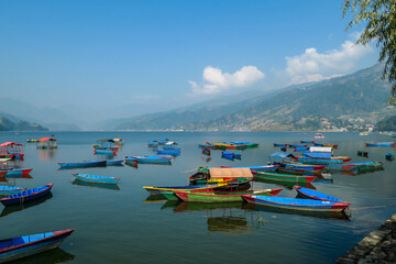 A view on Phewa Lake in Pokhara, Nepal with many colorful boats anchored around the shore. There are high Himalayan ranges in the back. Calm surface of the lake. Clear and sunny day. Serenity