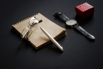 Glasses, a metal ballpoint pen, an eco-friendly notebook, a wristwatch and a red box lie on a black...