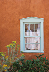 Window frame painted blue in red stucco plaster wall with tall yellow plants. Pueblo adobe building in Southwest USA