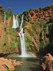 Ouzoud Waterfall in Morocco is an enclave of green and cooling water, popular spot for natives and a destination of one day tourist tours. Green vegetation,  red rock and white water, clear blue sky. 