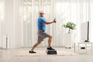 Mature man exercising on a step aerobic platform with dumbbells in front of tv