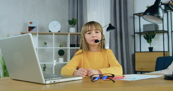 Cute satisfied smiling 10-aged girl in headset dressed in yellow sweater sitting in front of camera during video conference with school teacher