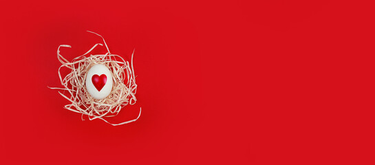 White egg with heart shape in decorative nest on red background with copy space.