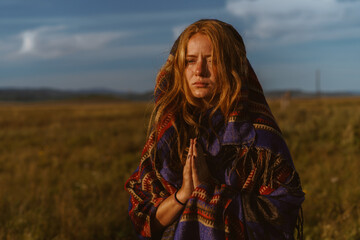 in a field among the grass against the sky and hills in the distance stands with a face expressing a plea and suffering wrapped in plaid girl prayerfully folded hands. High quality photo