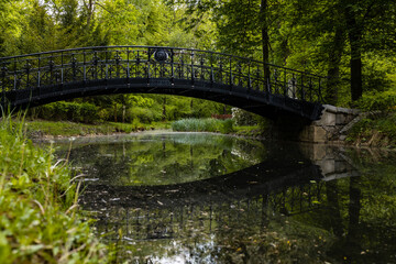 Fototapeta na wymiar Old wooden bridge with metal railing reflected in silent pond in park