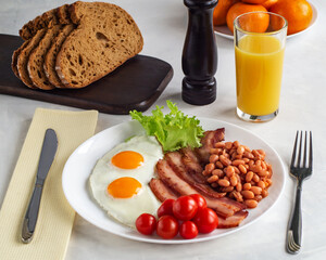Hearty breakfast with scrambled eggs, bacon, beans, cherry tomatoes, homemade bread, orange juice on a concrete countertop. English breakfast concept.