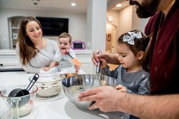 Dad helping toddler daughter cook while mom plays with baby in kitchen