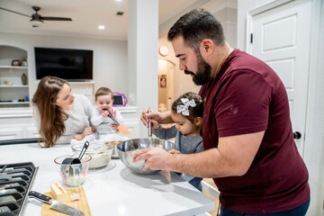 Dad helping toddler daughter cook while mom plays with baby in kitchen
