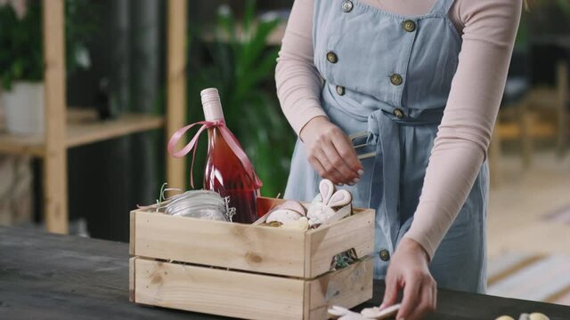 Tilt down shot of cheerful young woman putting Easter cookies and jar of dyed quail eggs into wooden box