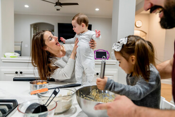Mom holding happy baby standing on counter while dad and daughter cook together