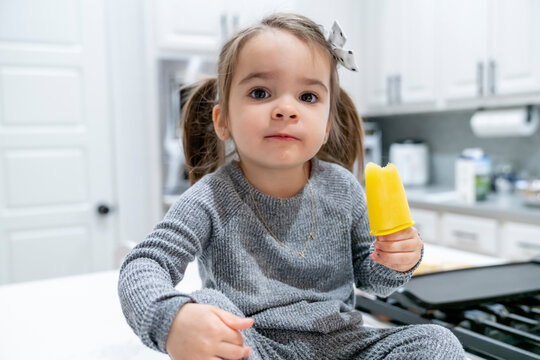 Girl sitting on kitchen counter eating orange popsicle