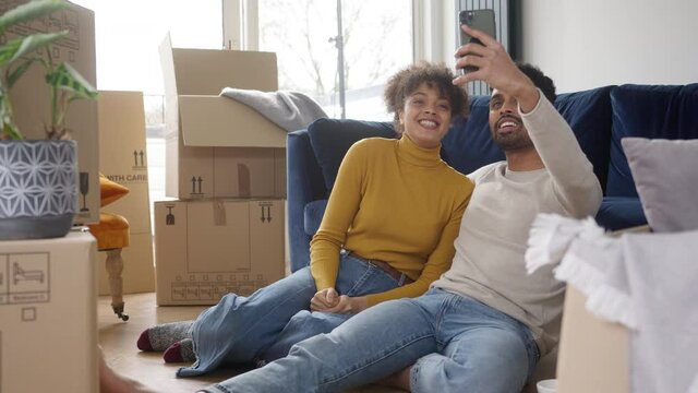 Young couple sitting on floor in lounge taking selfie on mobile phone with unpacked boxes in new home on moving day - shot in slow motion
