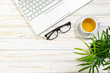 Minimalistic composition with cup of herbal tea, laptop, glasses and green flower in a pot on light wooden background. Office desktop concept. Cop space for your design. Selective focus.