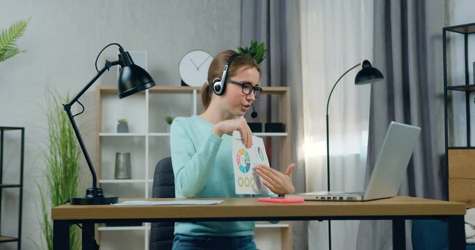 Pretty positive confident 20-aged female student in headphones presenting her report with infographics to university teacher during online seminar,remote education concept