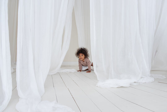 Little curly African American girl has fun laughs dances among airy white tulles fabric. Minimalist interior, Scandinavian style. Happy childhood. The concept of adopted children. Mixed ethnicity race