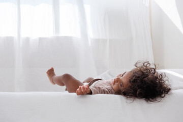Little curly African American girl lies on a white bed, has fun, laughs, smiles, plays. The concept...
