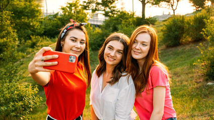 Three young girl friends taking picture of themselves on cell phone at summer sunny day. Outdoor portrait of three friends taking photos with smartphone