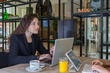Mujer de negocios caucásica trabaja en ordenador portátil en mesa cafetería y mira a otra persona frente a ella, quien trabaja en tablet