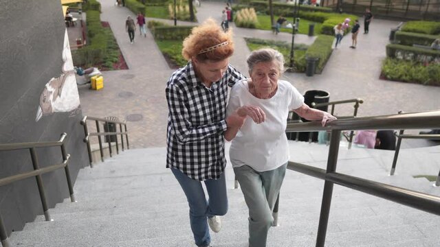 Home carer helping disabled woman getting down the stairs. Nurse helping a senior woman walk on stairs. Caregiver helping retired people go on stairs outside. Providing help and support for elderly