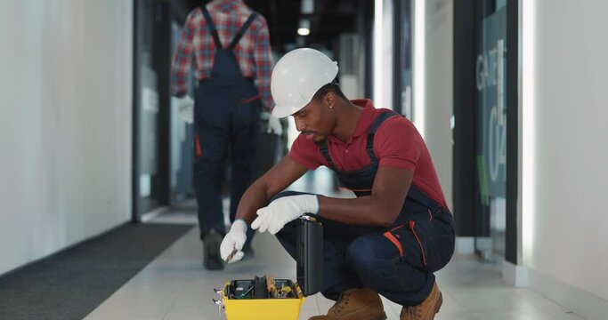 Afro-american young engineer architect wearing protective bodysuit searching right tool to repair electrical lighting system in building.