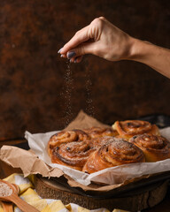 Woman's hand sprinkles sugar on freshly baked homemade cinnamon buns on brown background. Homemade...
