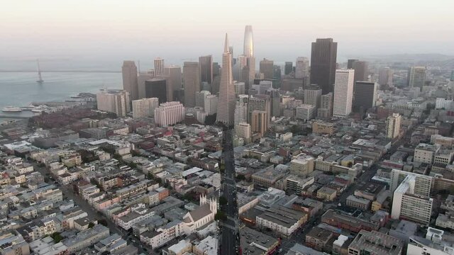 Aerial moving forward towards amazing downtown skyline under a hazy sky with the Oakland Bay Bridge in the background and cars bustling along busy city streets - San Francisco, California