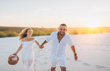 A young bearded man and a blond woman run and hold hands on the white sand at sunset. Love in the desert newlyweds. The love story of fun young people.