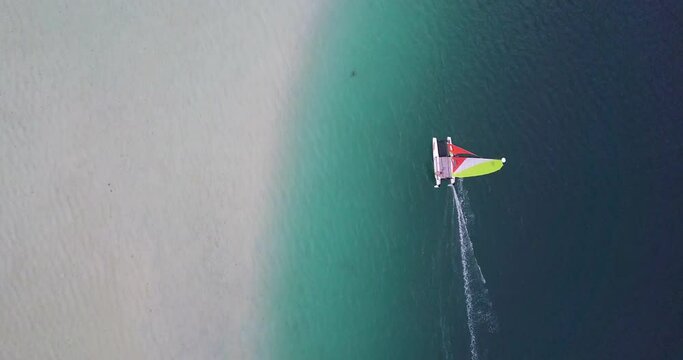 Aerial tracking over a windsurfer sailing across topical waters with brightly lit sand, gentle waves, and a turquoise colored sea - Oahu, Hawaii